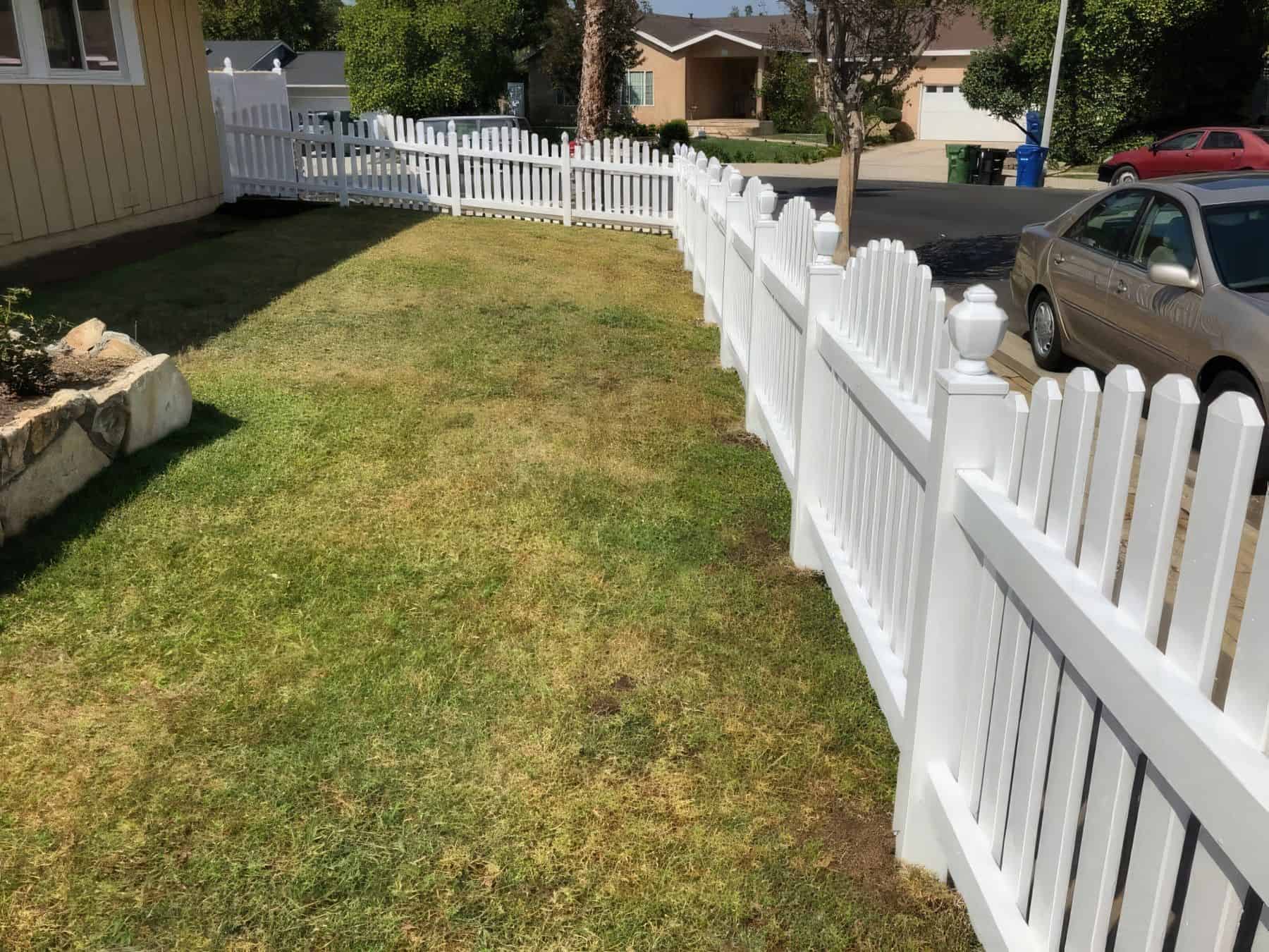Vinyl dog ear picket fence & ranch suburban house, with cars in the background. Concrete sidewalk & driveway complete the scene.