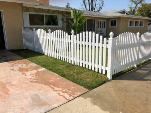 Vinyl dog ear picket fence in front of a ranch suburban house, with concrete sidewalk & driveway, and trees in the background.