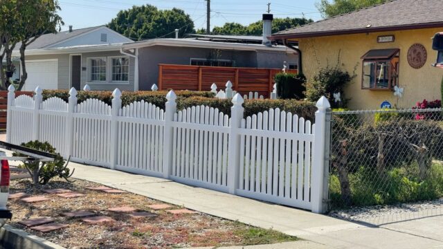 Suburban house with vinyl dog ear picket fence, concrete sidewalk, and green front lawn.