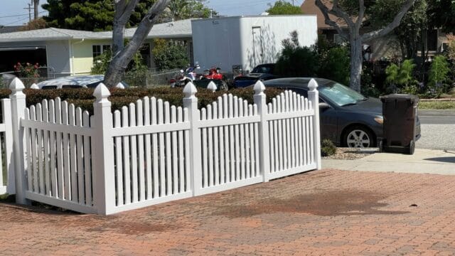 Vinyl dog ear picket fence & brick driveway surround suburban house with concrete sidewalk & front lawn.