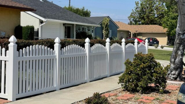 Vinyl dog ear picket fence surrounding suburban house with concrete sidewalk and neat front lawn.