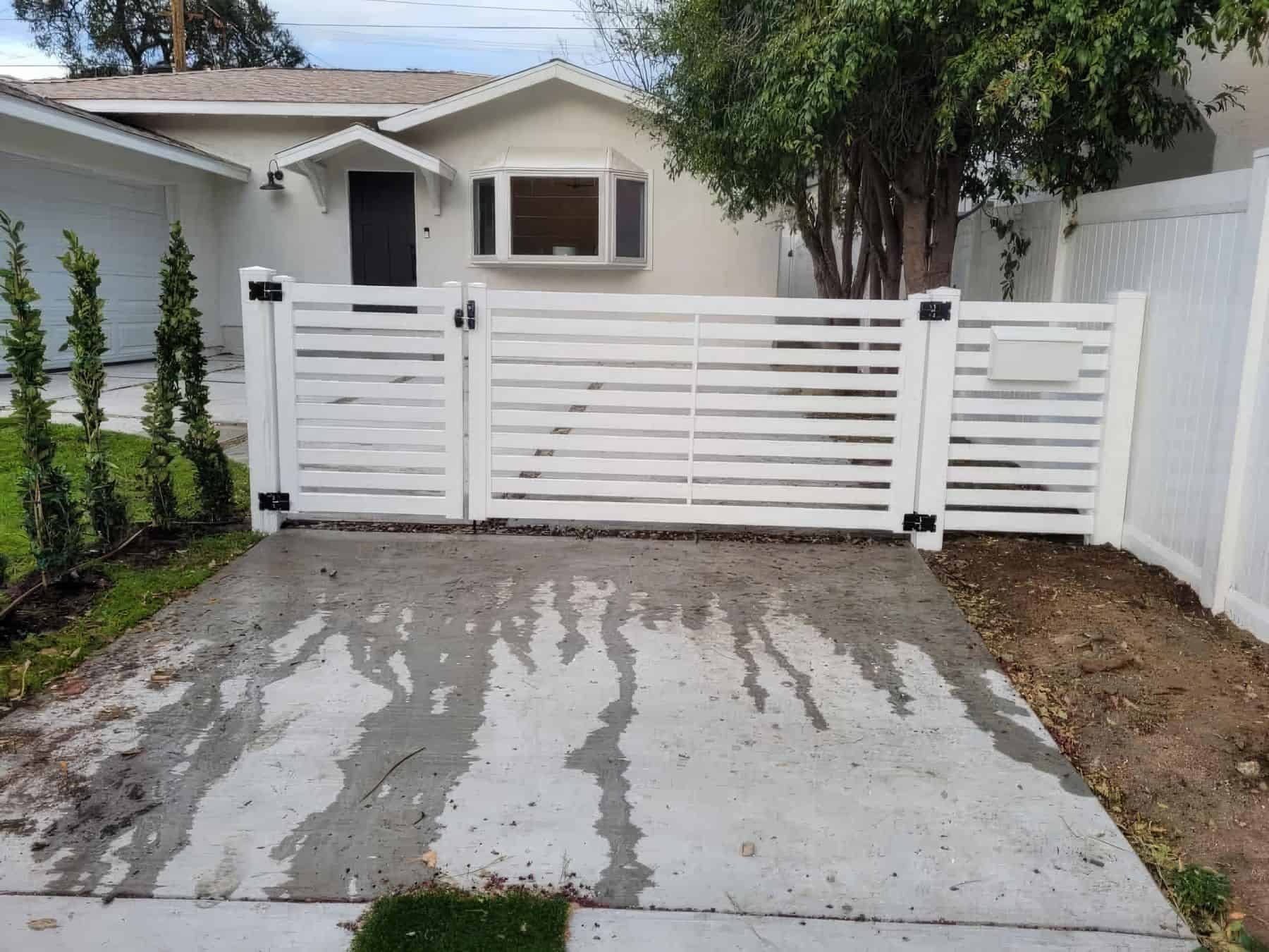 Vinyl fence gate leading from concrete driveway into charming suburban home surrounded by trees with a grassy front lawn.