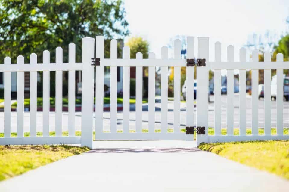Vinyl fence gate opening to concrete sidewalk, bordered by lush grassy lawn, with beautiful trees in the background.