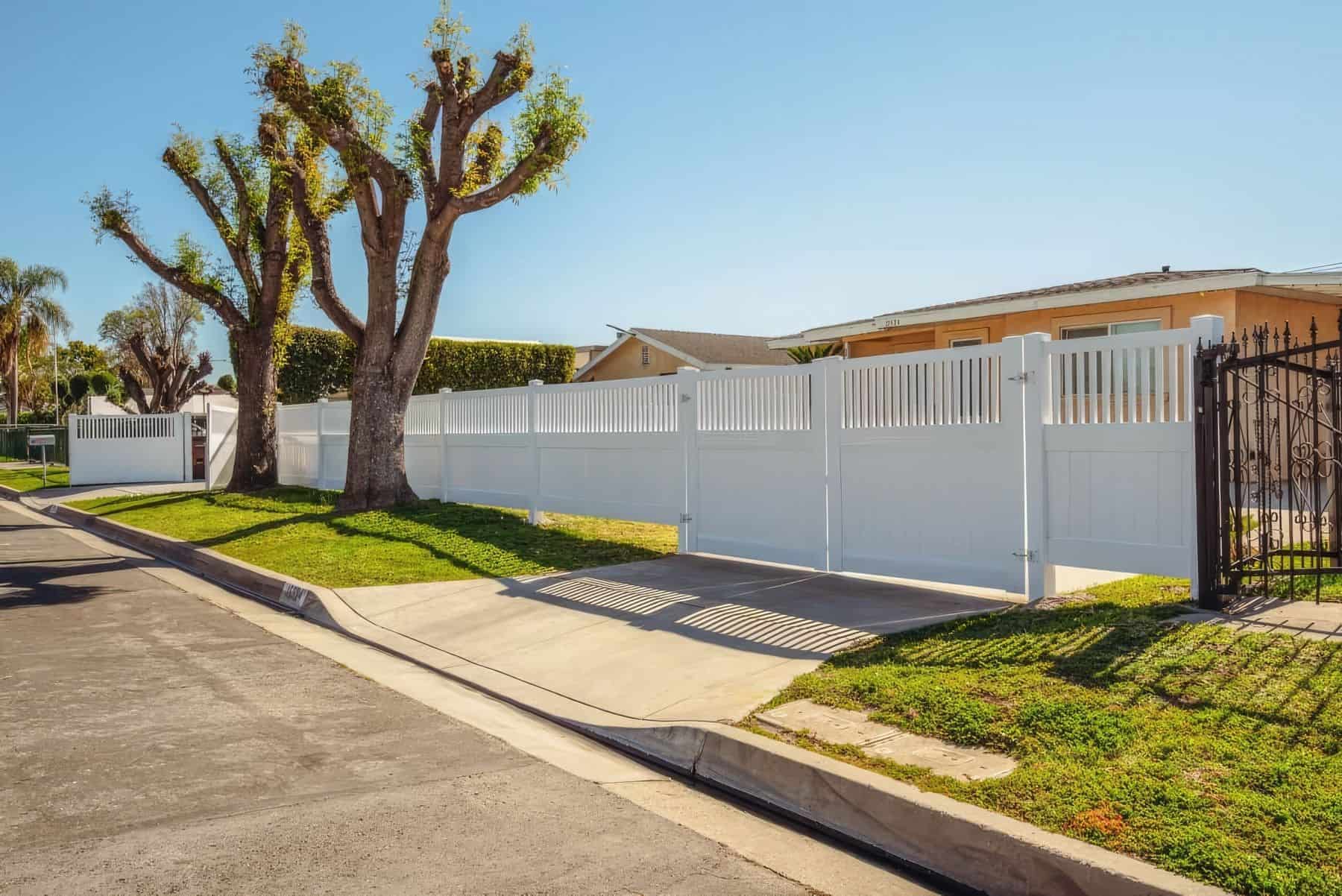 Vinyl fence gate & concrete sidewalk leading to a lush grassy lawn with trees in the background. Serene outdoor scene.