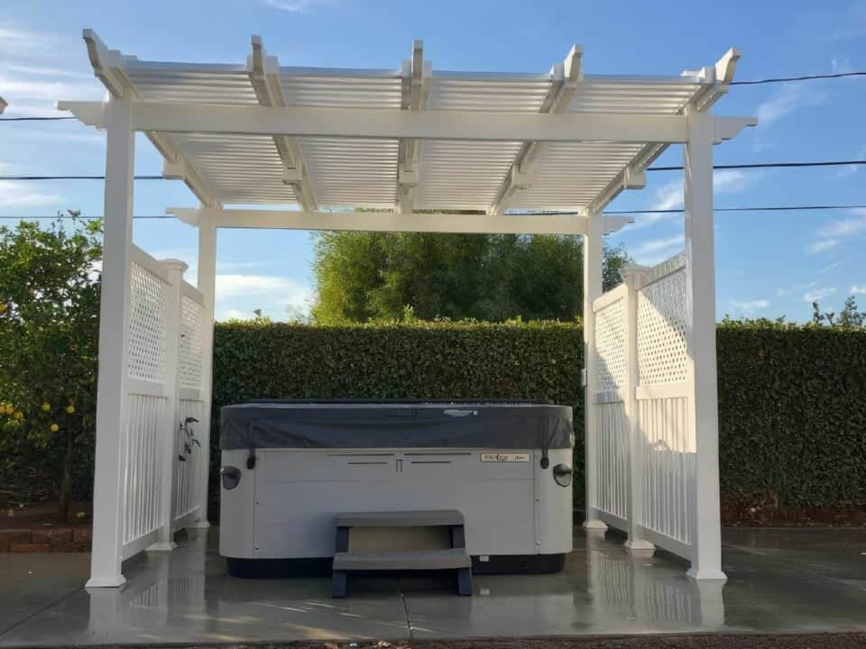 Vinyl free standing patio cover over concrete floor, jacuzzi, lush grassy lawn, and trees in the background.