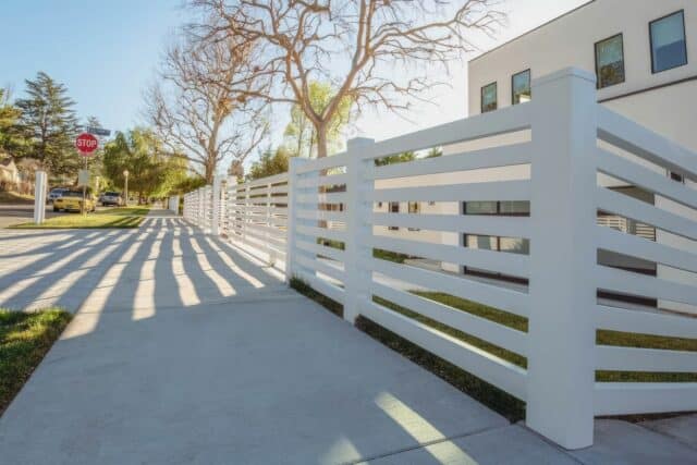 Vinyl horizontal picket fence lines concrete sidewalk, framed by lush trees in background, alongside a serene grassy lawn