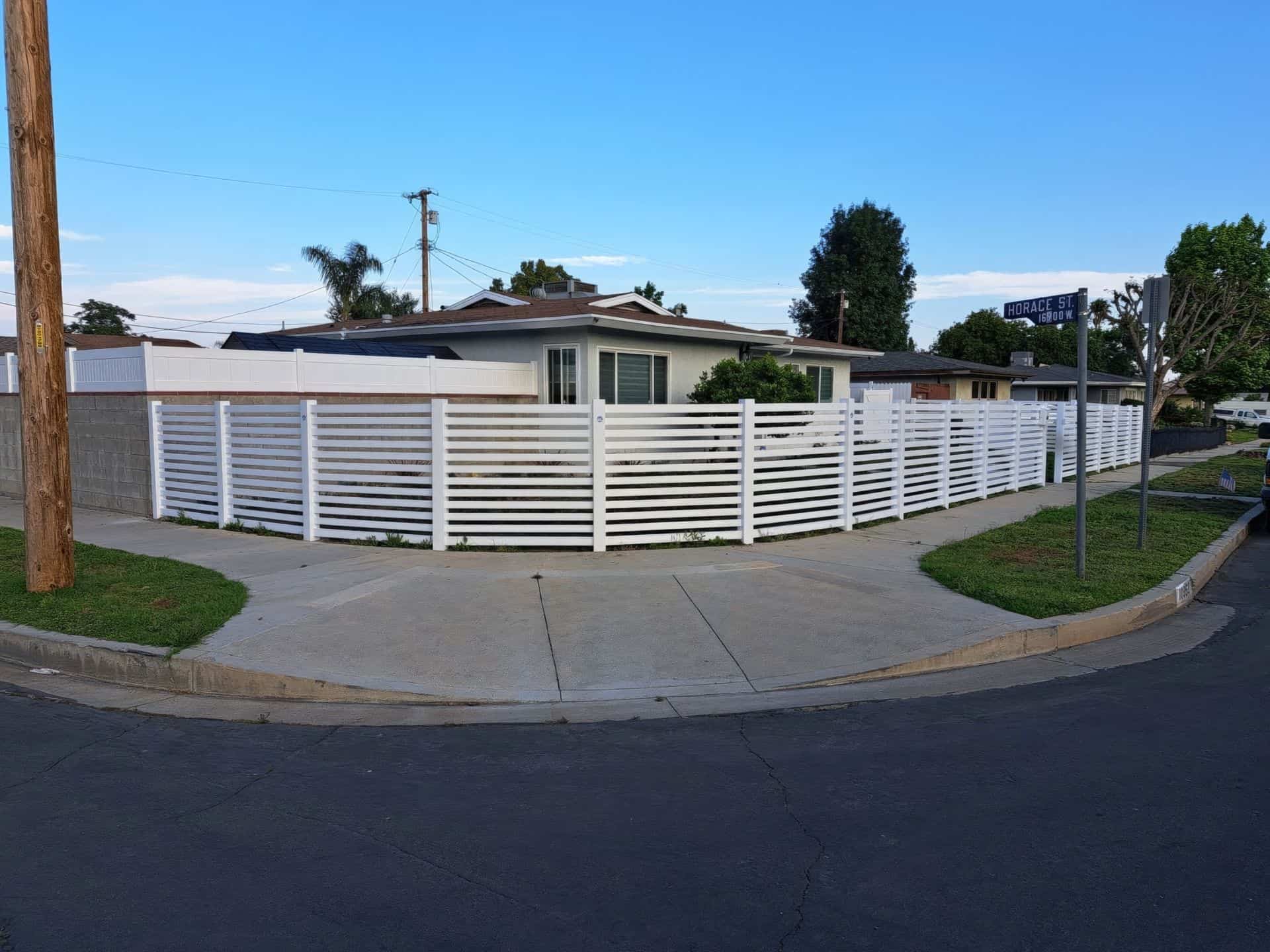 Vinyl horizontal picket fence & suburban home with concrete sidewalk, lush lawn, & trees in the background.