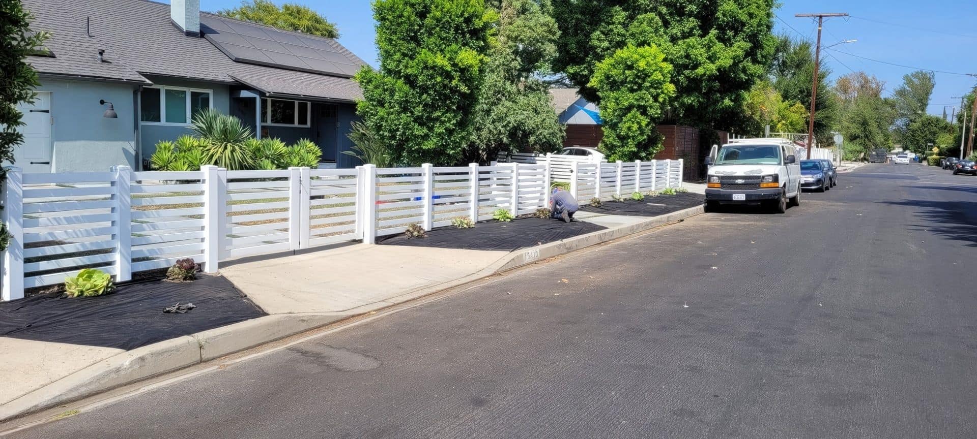 Suburban home with vinyl horizontal picket fence, concrete sidewalk, lush lawn & trees in the background.