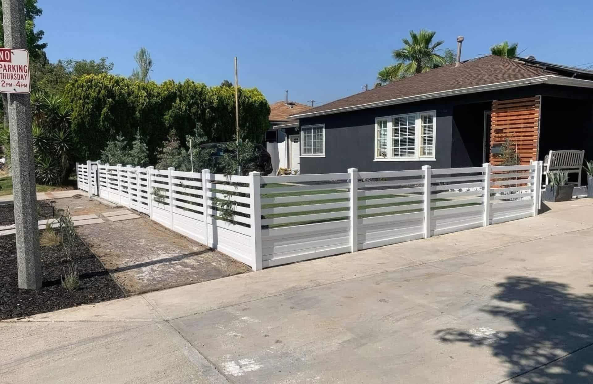 Vinyl horizontal picket fence lining concrete sidewalk & driveway, with lush front lawn & distant trees in background.