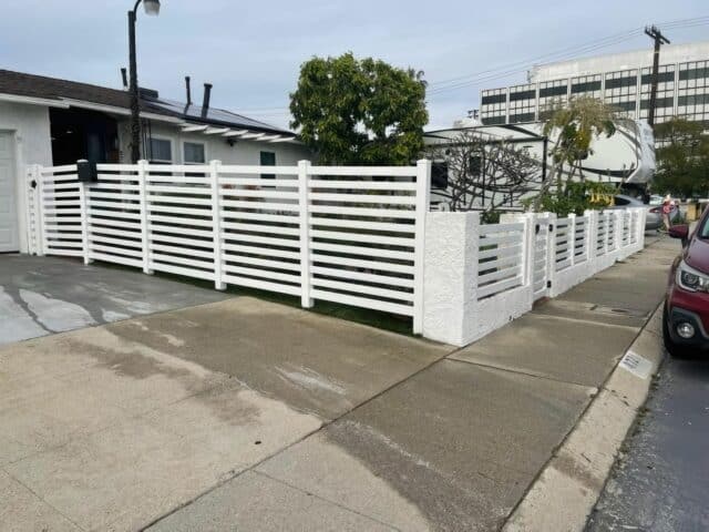 Vinyl horizontal picket fence alongside concrete sidewalk & driveway, front lawn, with trees in background.