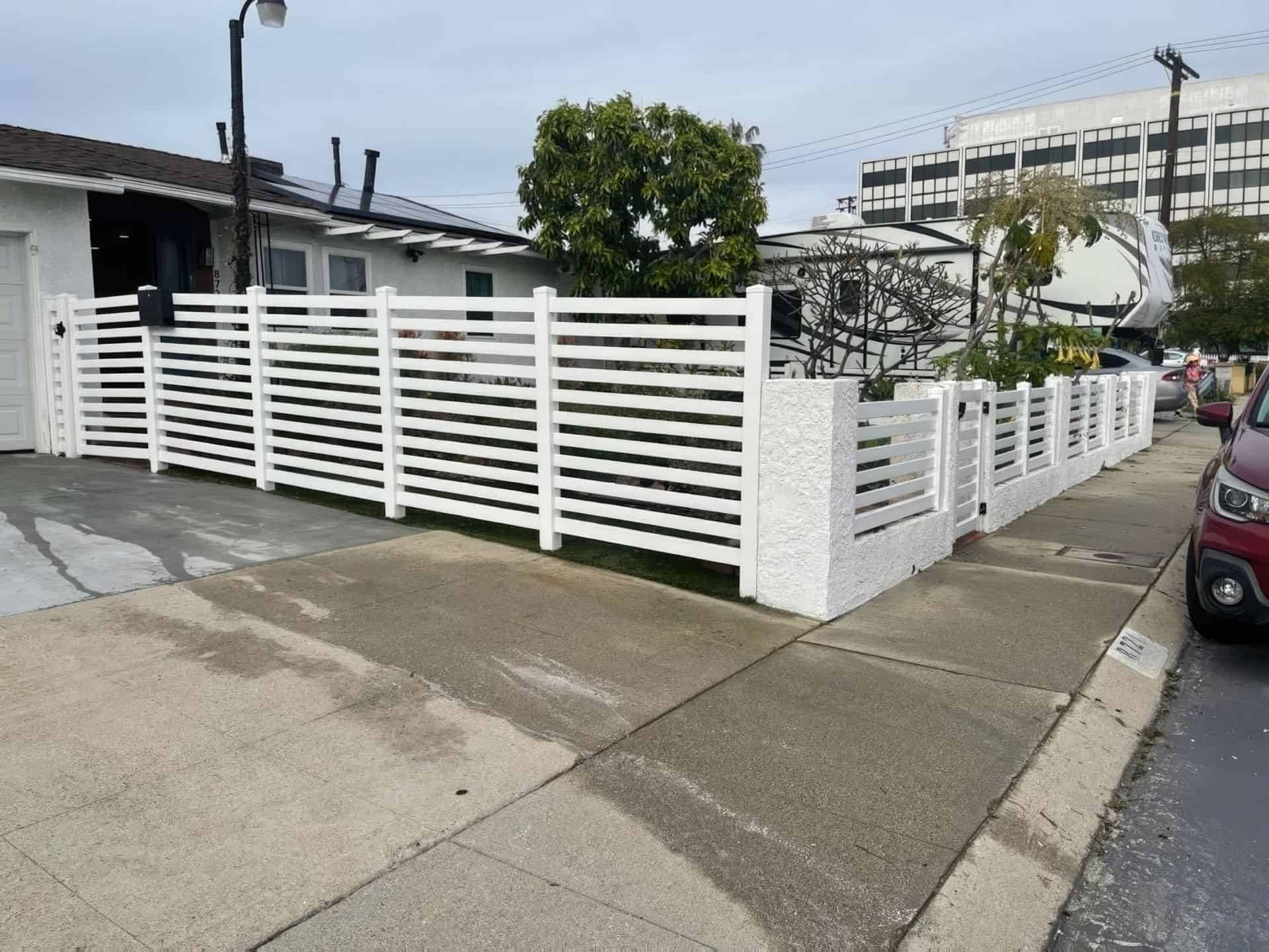 Vinyl horizontal picket fence alongside concrete sidewalk & driveway, front lawn, with trees in background.