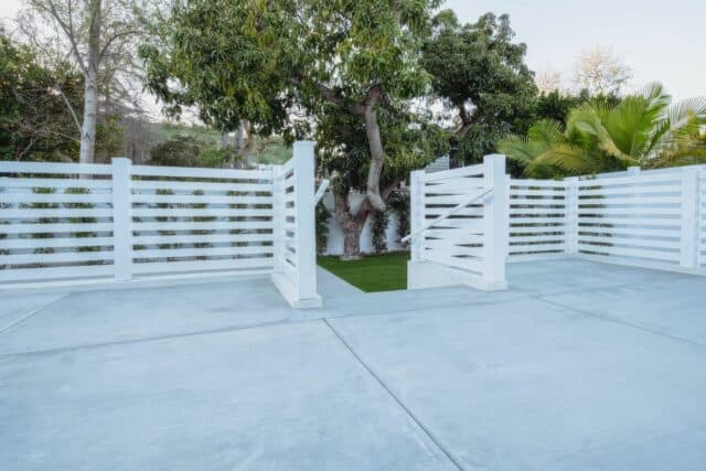 Vinyl horizontal picket fence by poolside, concrete floor, small garden, lush grass lawn, trees in background.