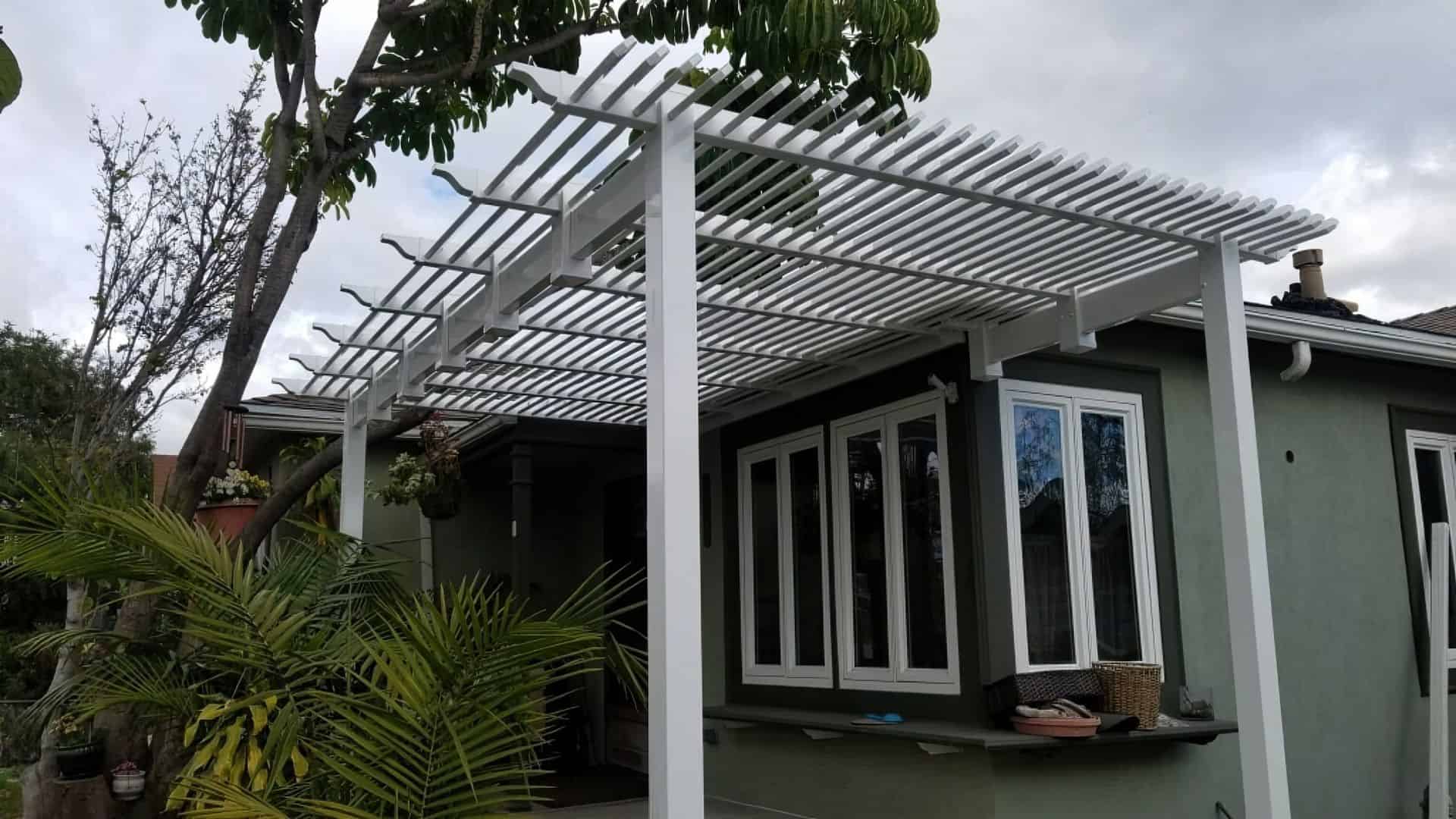 Vinyl louvered patio cover over small garden with potted plants and various small flowers leading to the grassy backyard