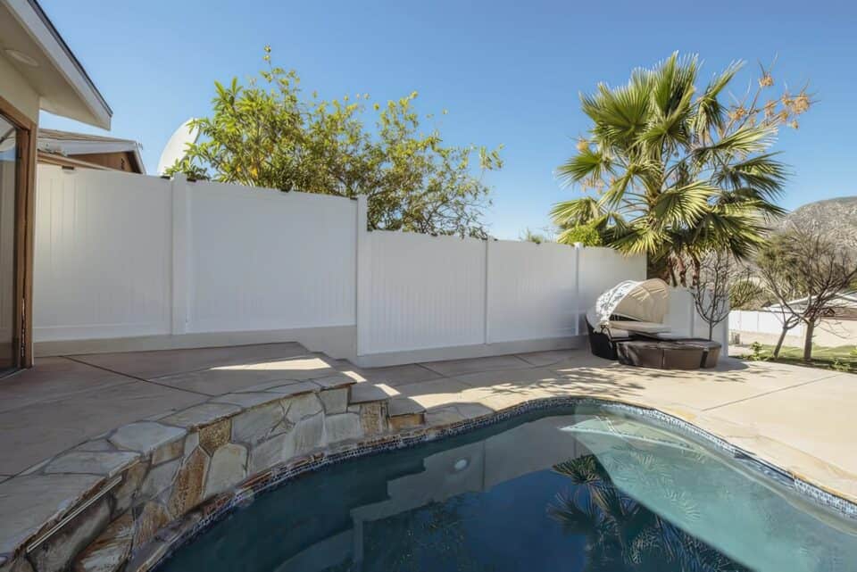 Vinyl pool fence with gate leading into area with concrete flooring and garden patch to the side with small trees.