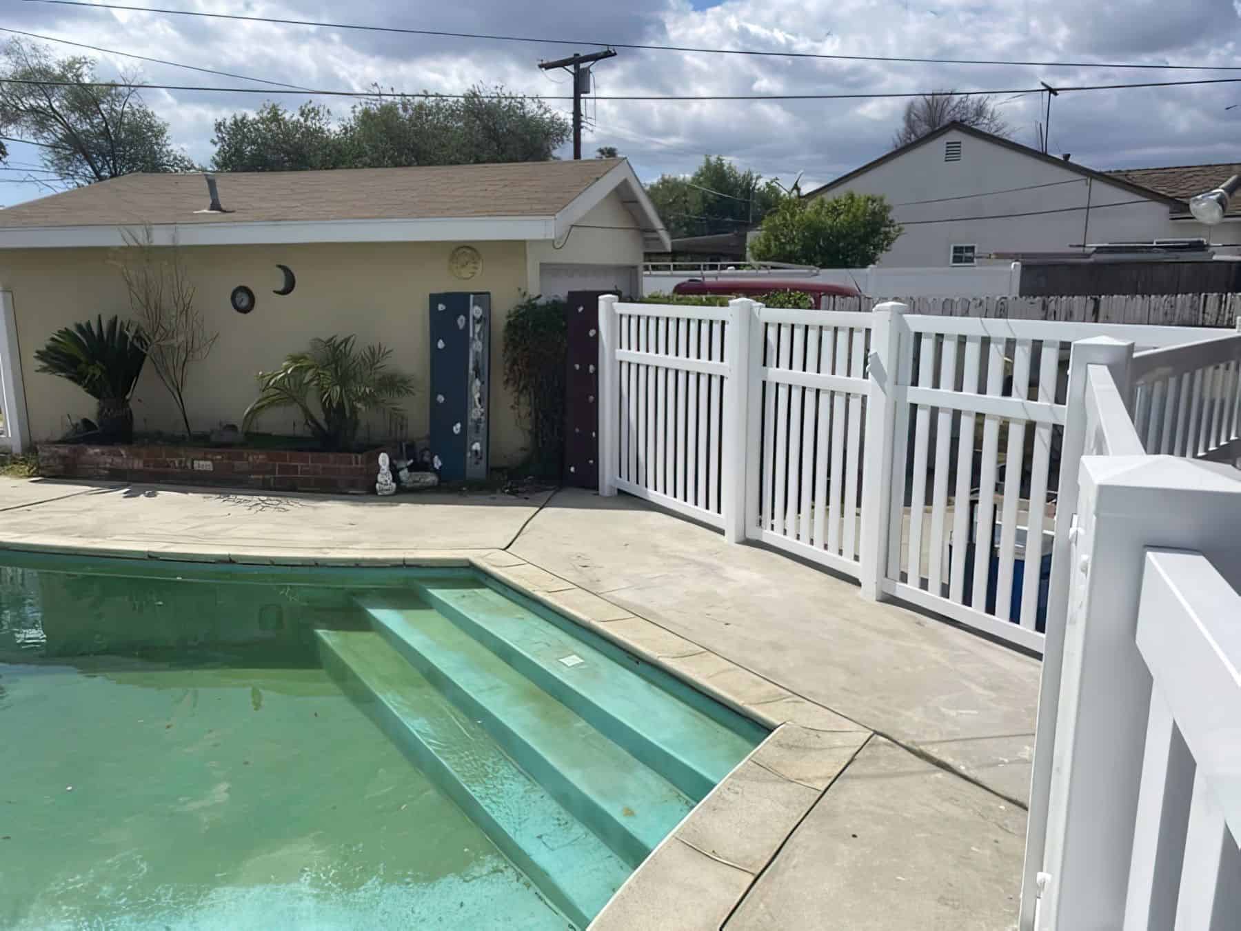 Vinyl pool fence behind small garden patch with palm trees and small flowers and uniquely shaped oval pool with stairs.