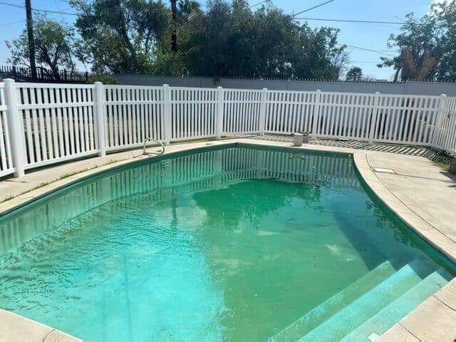 Vinyl pool fence with gate leading out to backyard beside the smaller suburban house with small garden patch and small trees