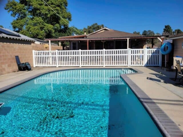 Vinyl pool fence on grassy lawn creating a boundary around the pool area with concrete flooring and trees in the background.