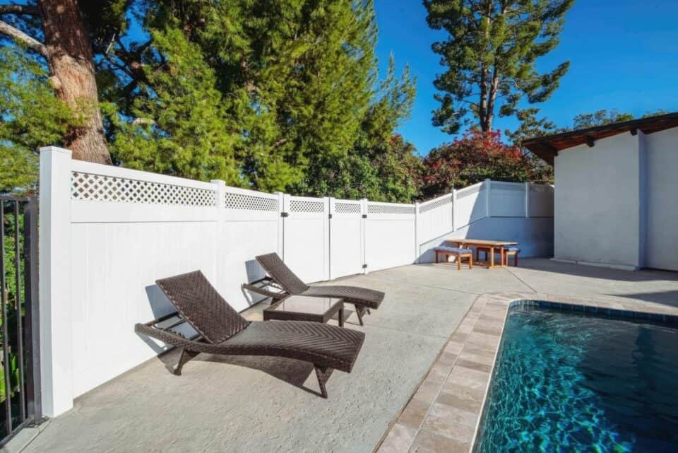 Vinyl privacy and lattice fence with gate leading into swimming pool backyard with relaxing chairs and concrete flooring.