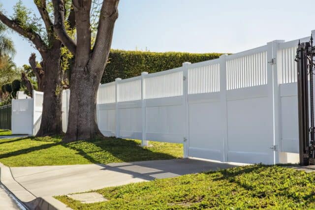 Vinyl privacy and picket fence with gate beside massive trees leading into driveway of suburban home