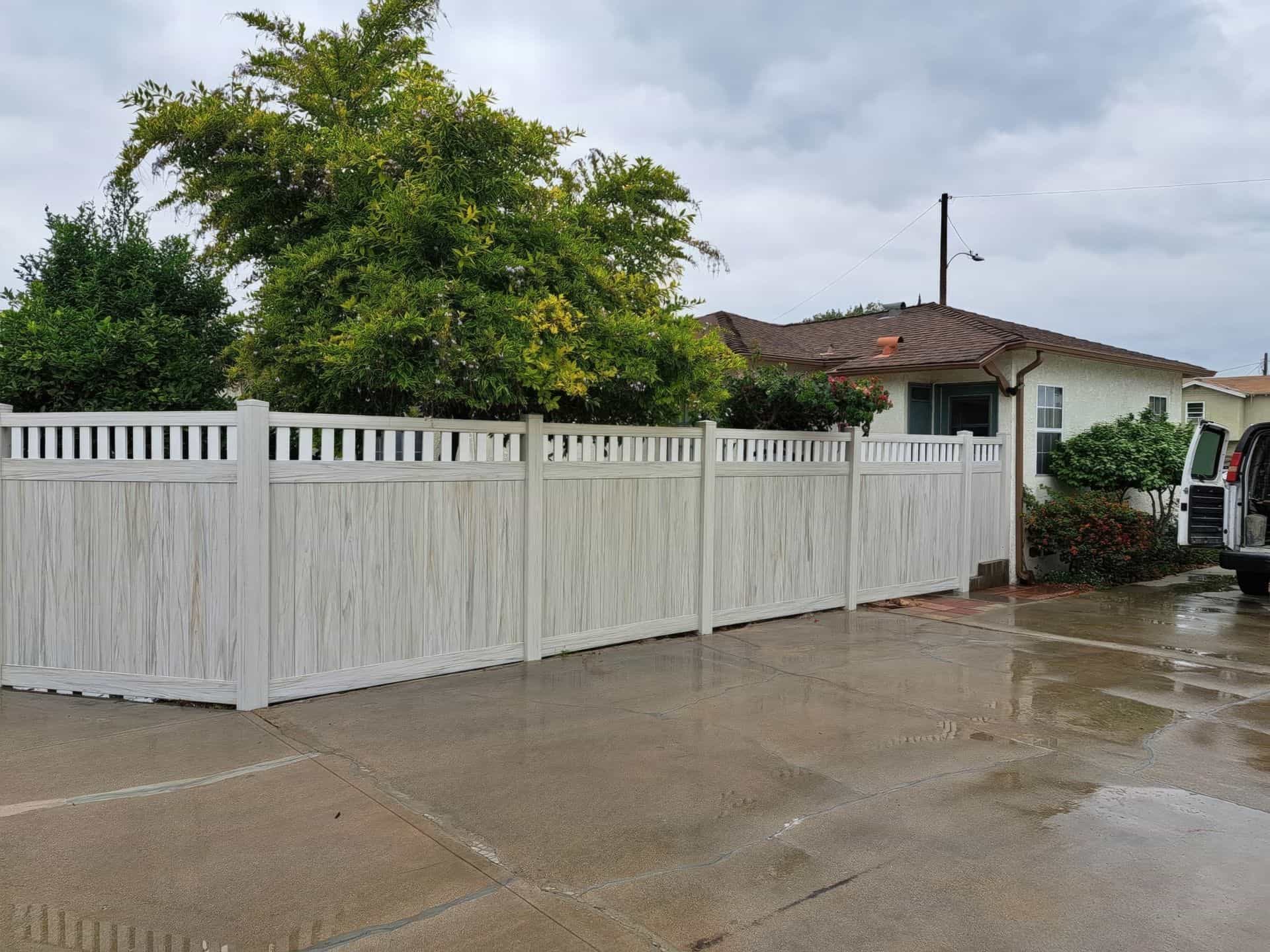 Vinyl privacy and picket fence separating concrete driveway from the other home’s garden featuring large trees and plants