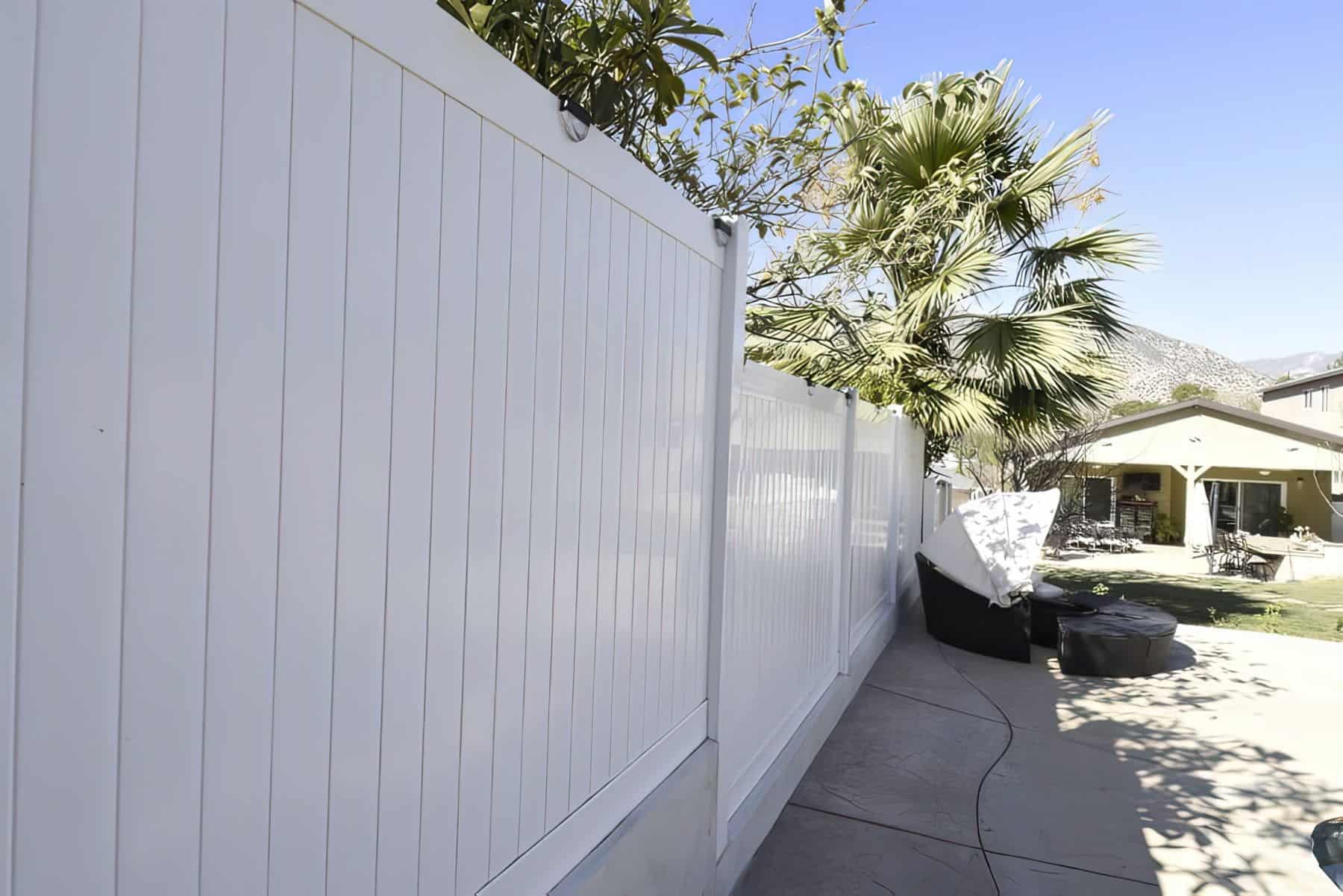 Vinyl privacy fence surrounding large concrete backyard with patches of grass and comfortable seating couches.