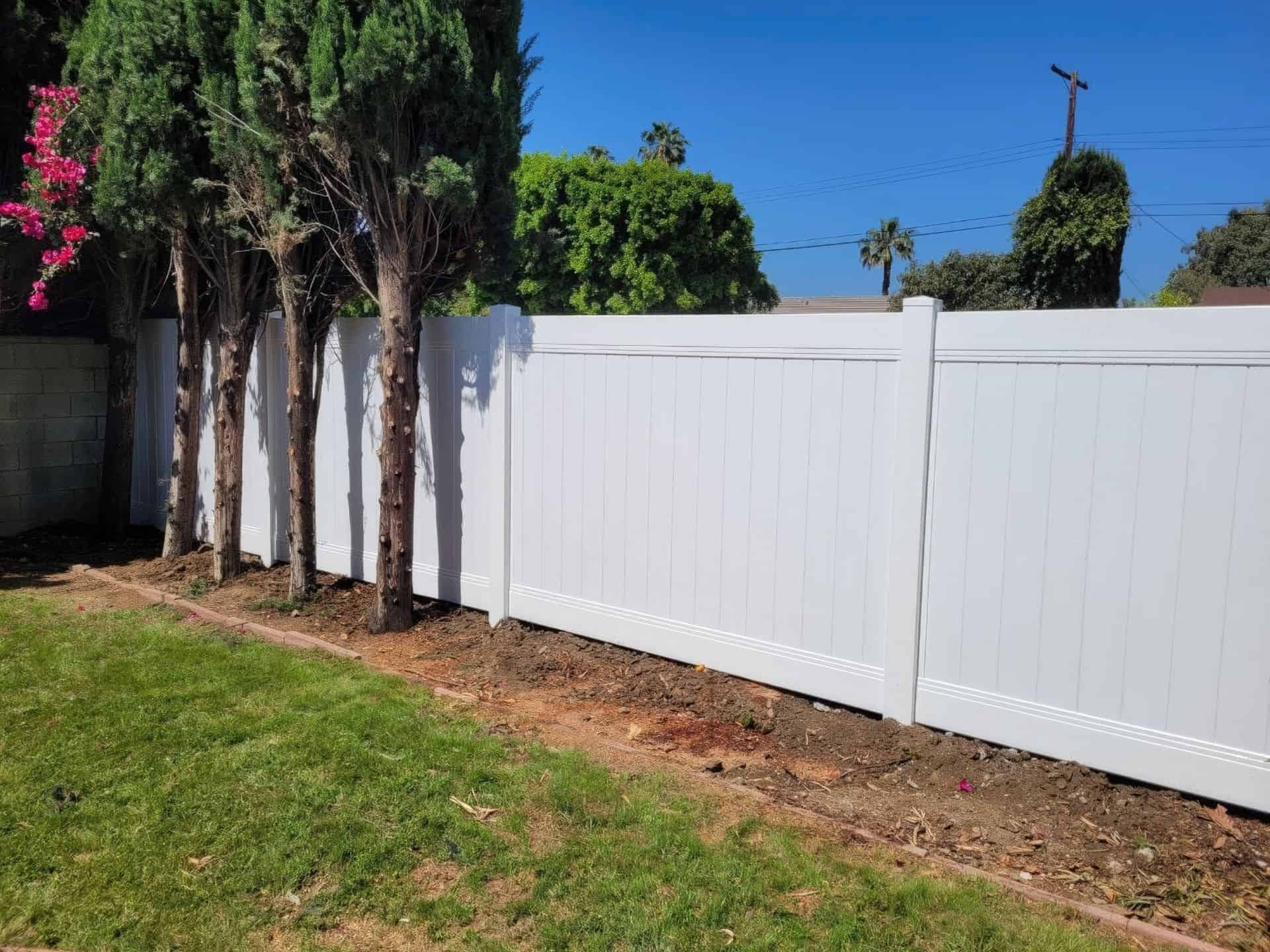 Vinyl privacy fence with behind small garden boundary with long trees at the corner of grassy lawn connecting to the house