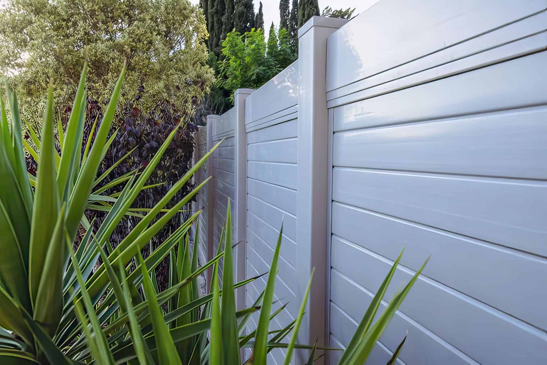 Matte vinyl privacy horizontal fence bordering the backyard, behind small trees and plants of garden patch.