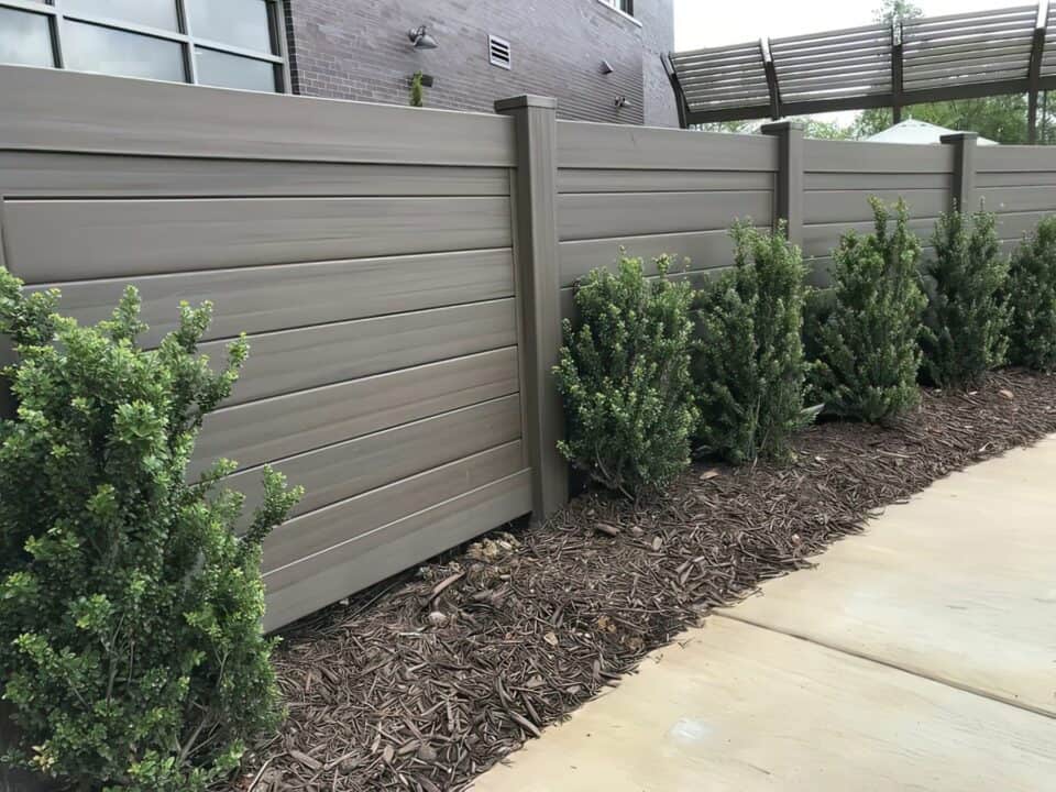 Rich brown vinyl privacy horizontal fence used as a boundary around the home with small plants separating the patio and sidewalk.