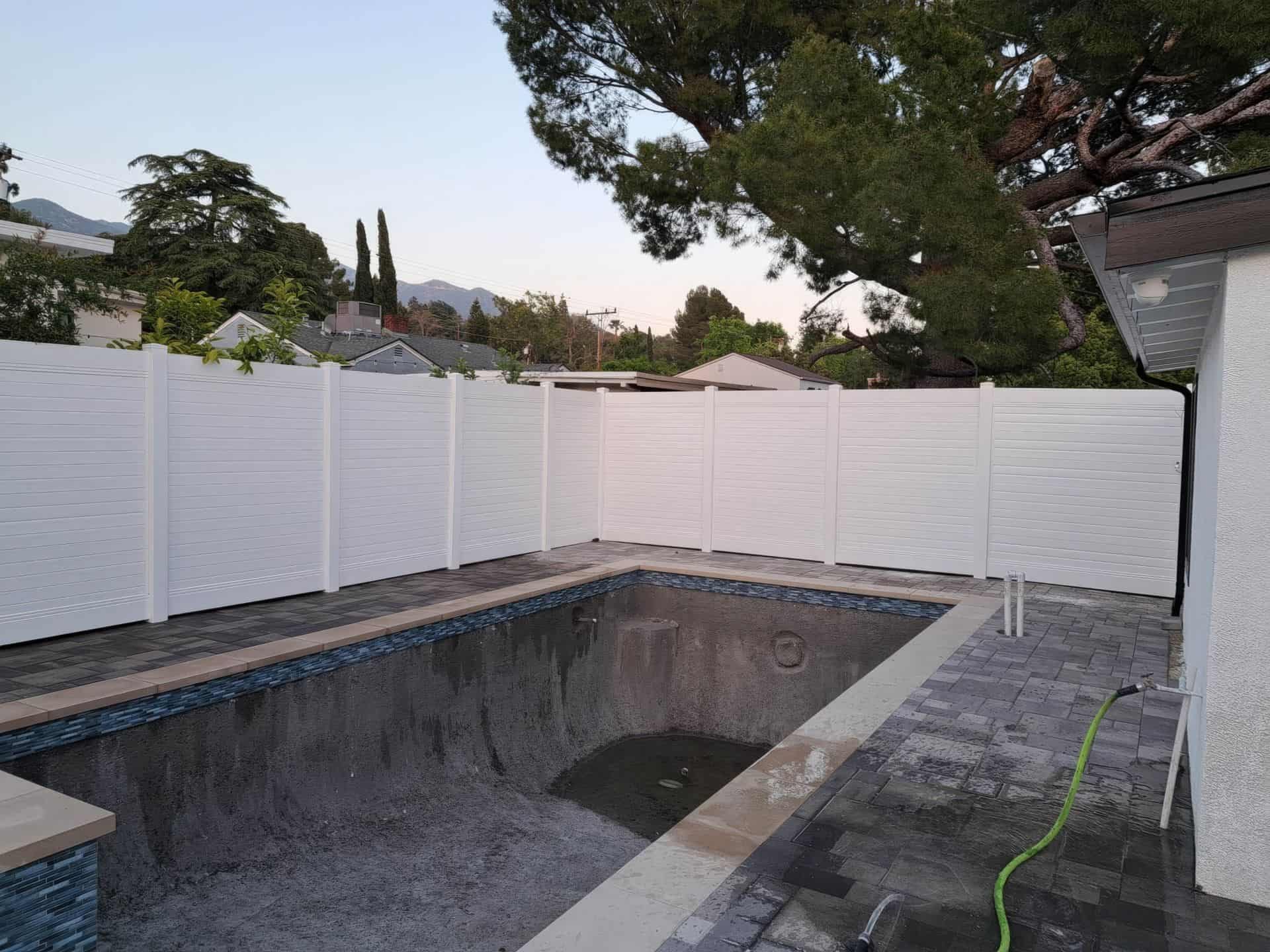 Vinyl privacy horizontal fence creating boundary around swimming pool with stylized concrete flooring and trees in the background.