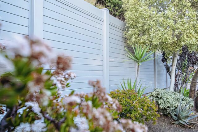 Vinyl privacy horizontal fence in front of lush backyard garden with multiple trees and small plants, along with a lush lawn