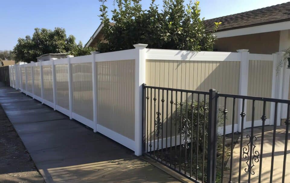 Vinyl privacy two-tone fence beside metal gate and concrete walkway with larger trees and a lush garden on the inside