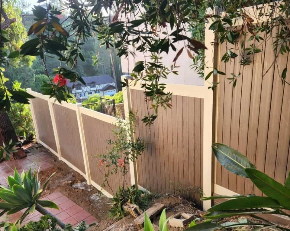 Vinyl privacy two-tone beige and brown fence in backyard with red tile flooring and multiple trees and small plants.