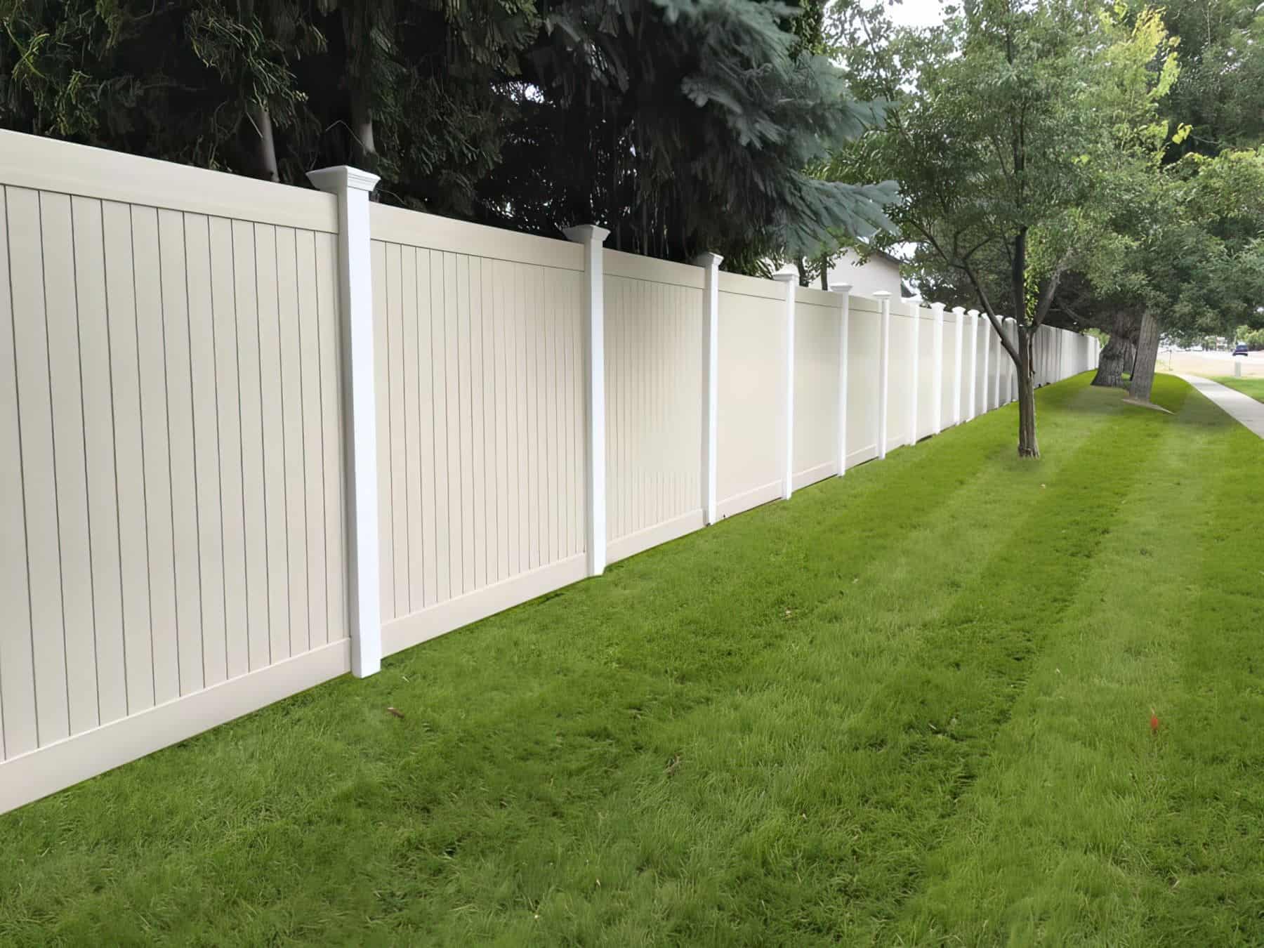 Vinyl privacy vertical fence as a boundary wall surrounded by trees and on top of well trimmed lush lawn.