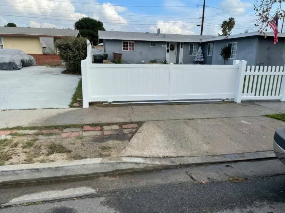 Vinyl rolling gate next to picket fence, behind concrete sidewalk, and in front of ranch style suburban house.