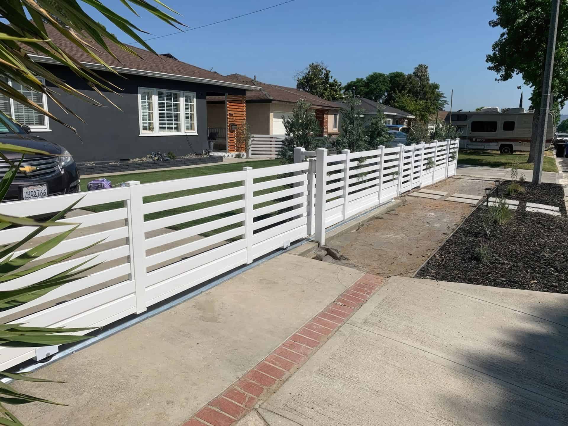 Vinyl rolling gate leading into the concrete driveway of ranch style suburban home with front lawn and trees in the back.