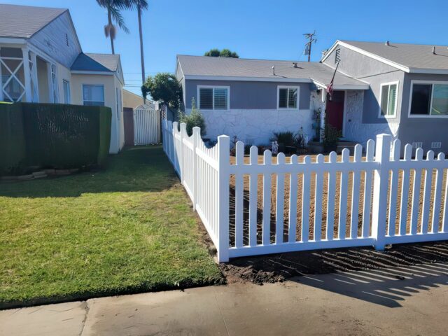 Vinyl scalloped picket fence separating two lawns of different houses surrounded by large hedges and small gardens.