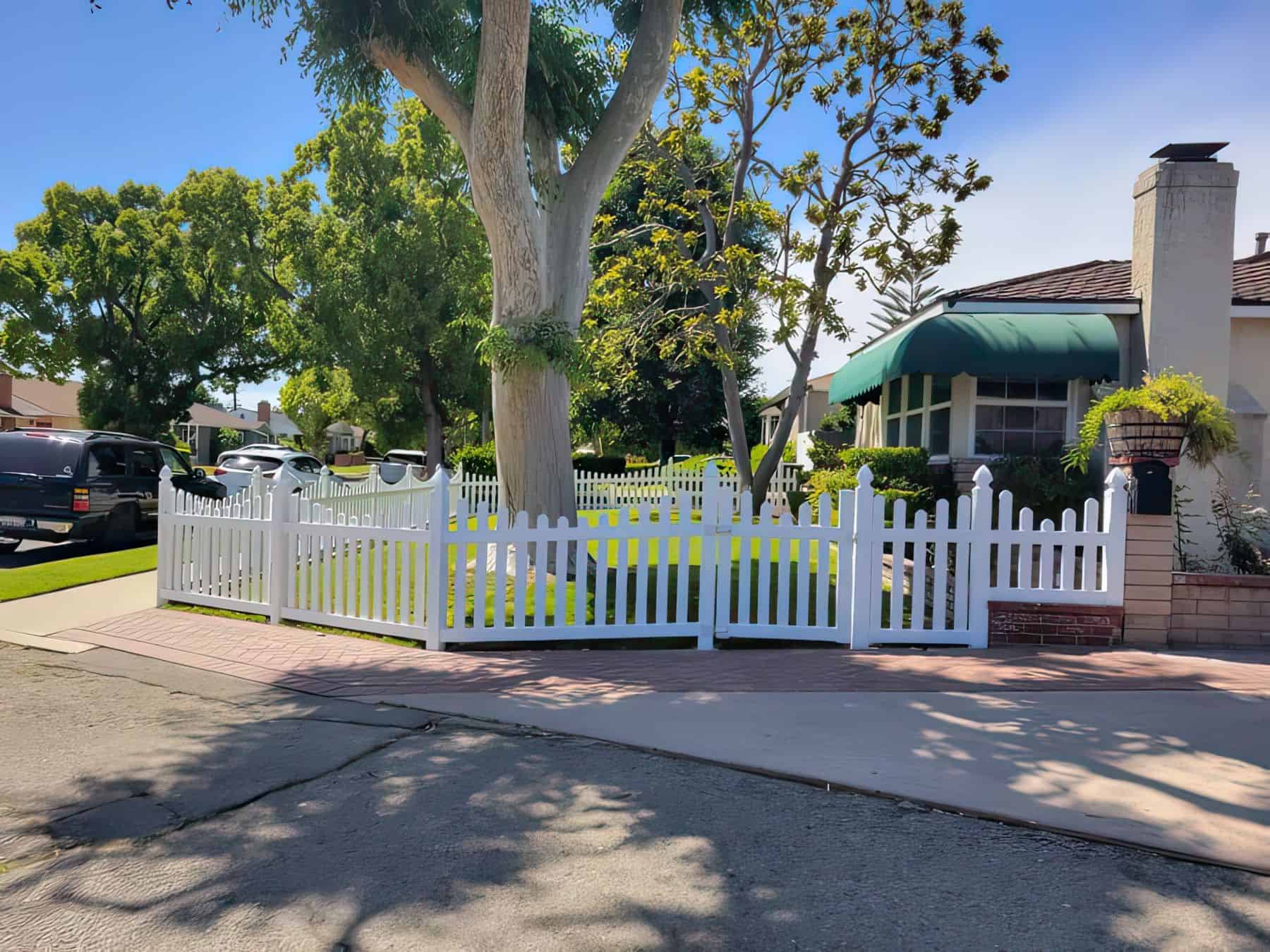 A cute vinyl scalloped picket fence creating a boundary around two big trees, a small lawn, and other potted plants
