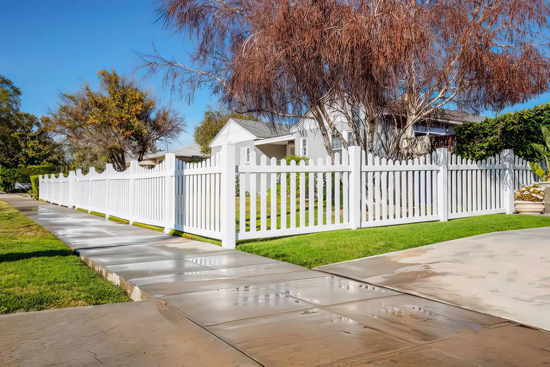 Fully enclosed vinyl scalloped picket fence surrounding a muted white colored home with large trees and a small garden.