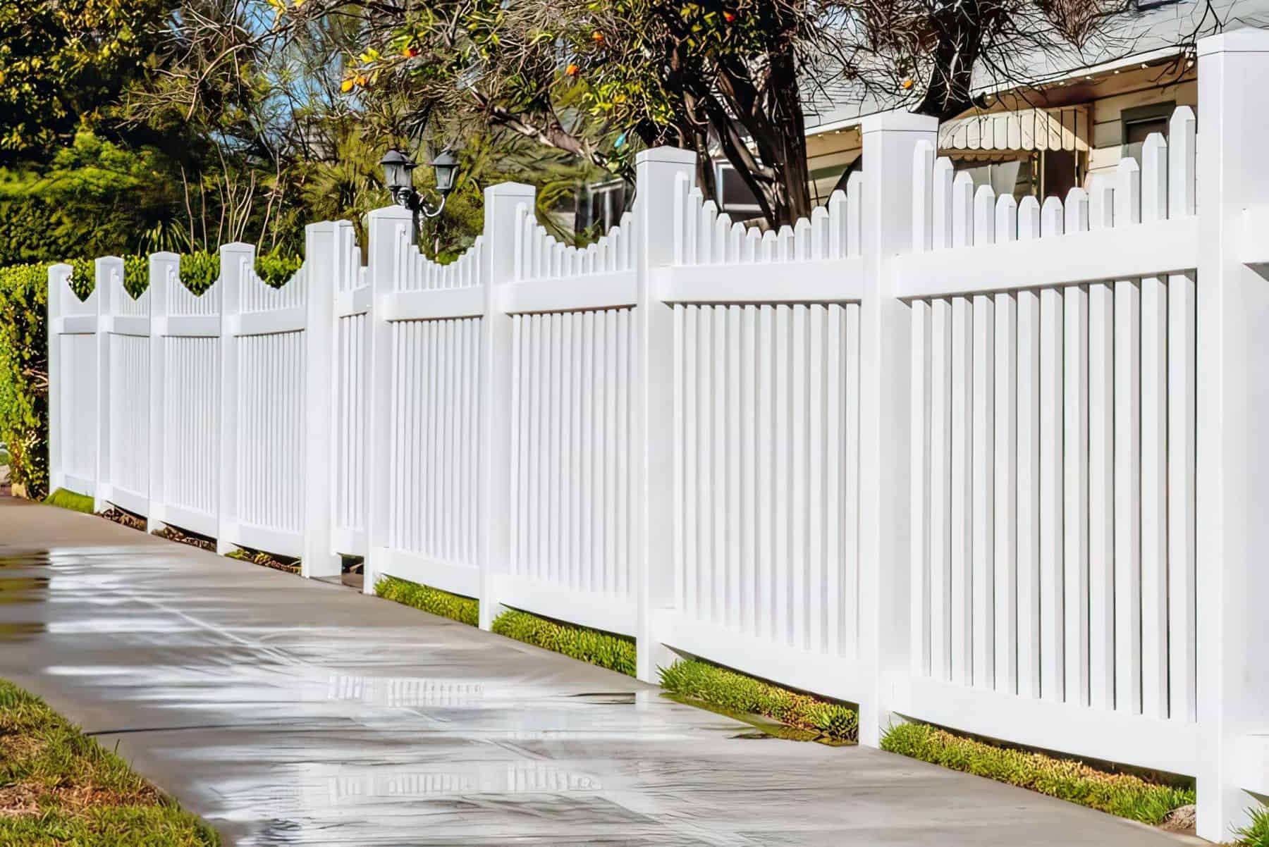 Pure white vinyl scalloped picket fence next to sidewalk and lush front lawn with trees and lampposts surrounding it.