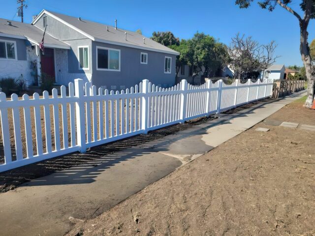 Vinyl scalloped picket fence with gate leading to entrance of small town home with a flag pole surrounded by trees.