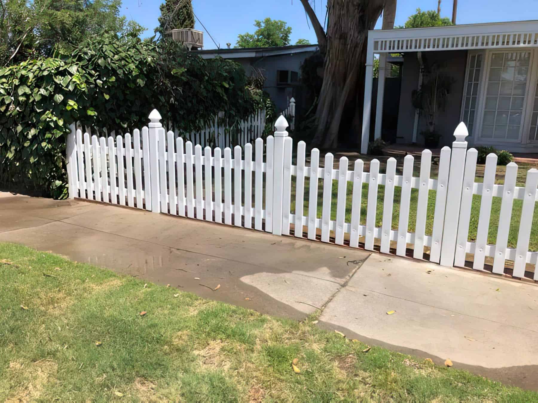 Vinyl scalloped picket fence in front of porch with a glass door entrance and a grassy pathway leading to the entrance.