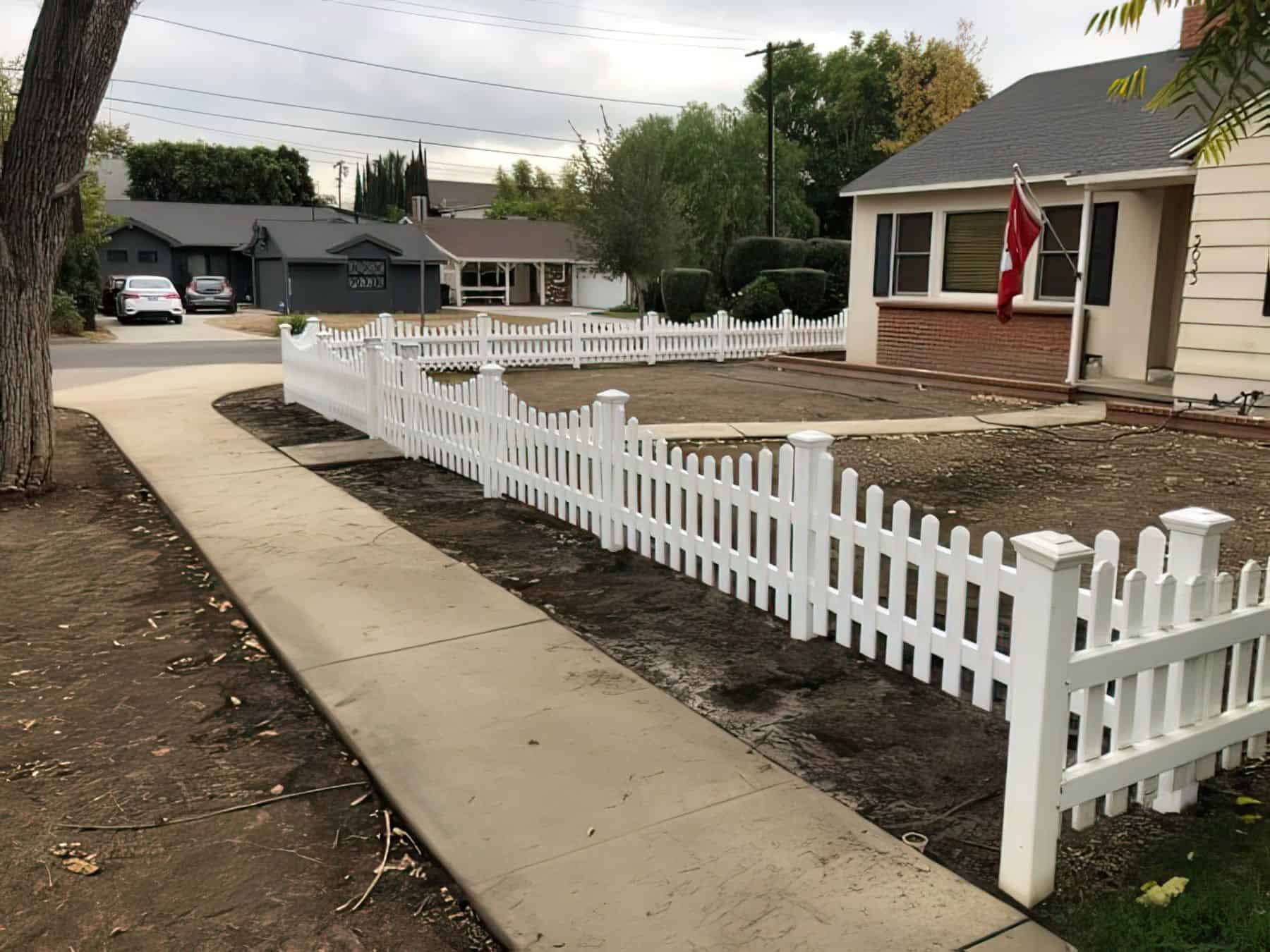 Knee high vinyl scalloped picket fence separating the sidewalk from the quaint suburban home with hoisted flag and trees.