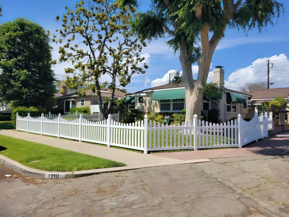 A short vinyl scalloped picket fence surrounding a ranch style home with a lush garden, big trees, and potted plants.