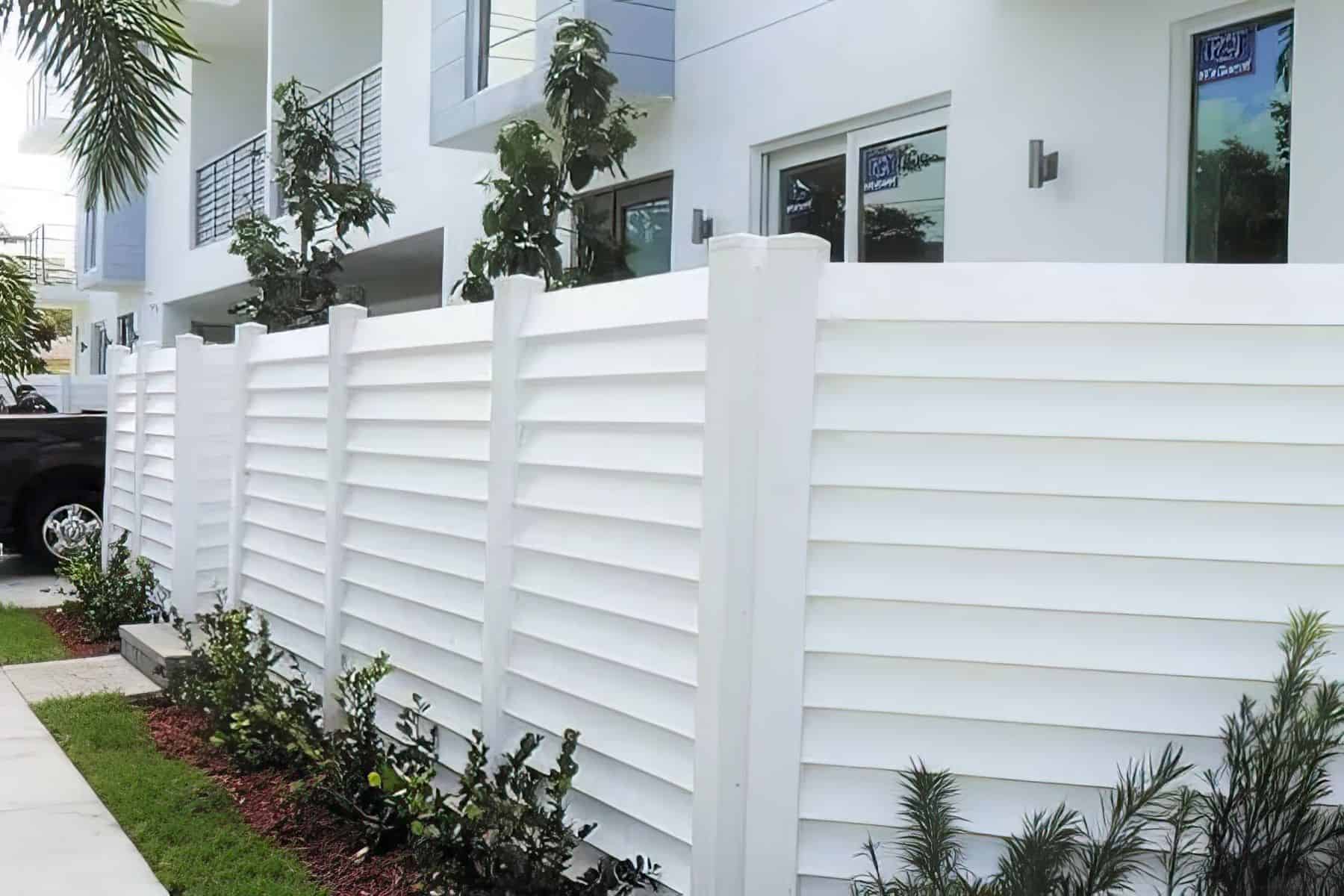 Louvered white vinyl semi-privacy fence separating the sidewalk and small patch of grass from the glass door entrance.