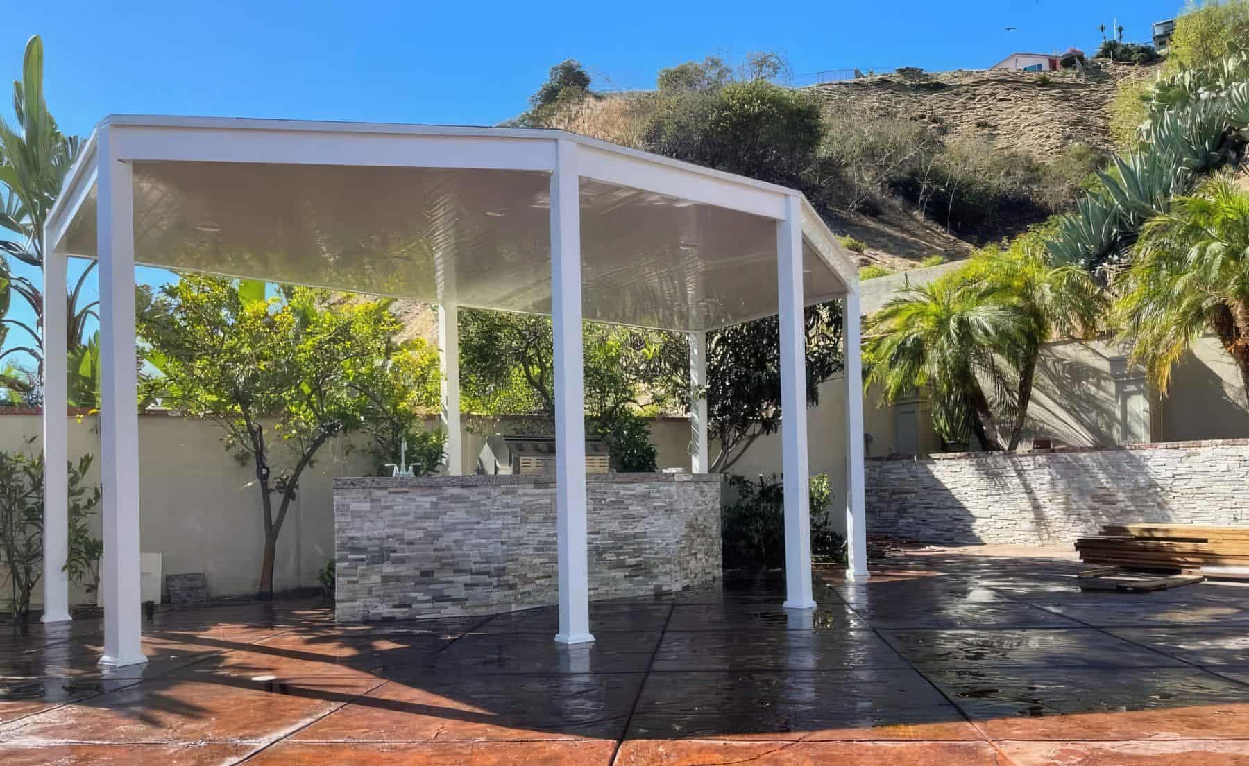 A vinyl solid patio cover in backyard escape surrounded by lush trees with a marble and granite kitchen top inside.