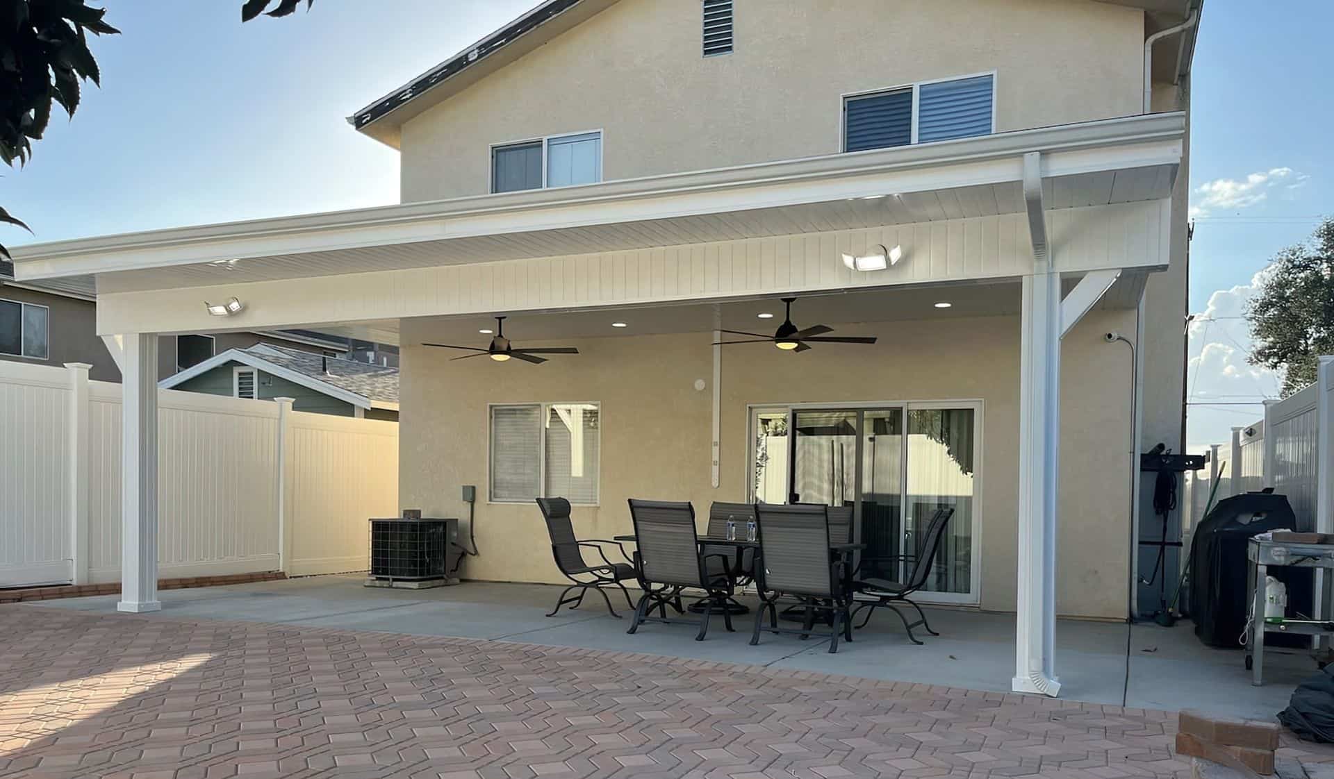 Chic vinyl solid patio cover with fans in the lush front yard with cozy outdoor furniture, and sleek tile flooring.