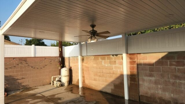 A vinyl solid patio cover with a ceiling fan in the backyard surrounded by wall offering relaxation in sunny weather.