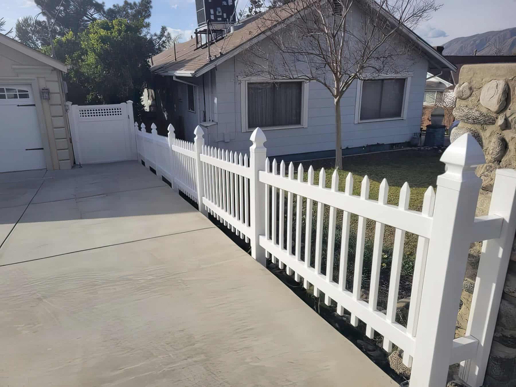 White vinyl vertical picket fence adjacent to vinyl gate, surrounding a lawn of a house with green grass.