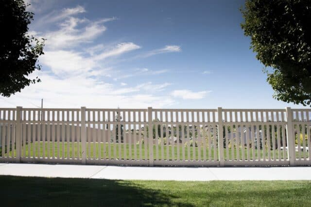 Vinyl vertical picket fence embraced by lush trees, set against a clear sky and vibrant green grass besides the pavement.