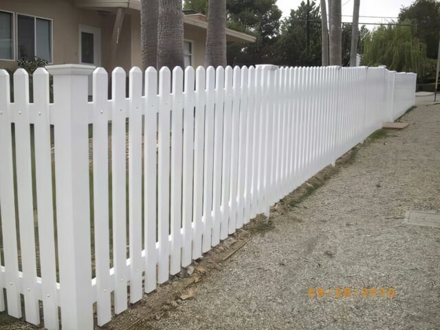 A vinyl vertical picket fence, adjacent to pavement, elegantly frames tall palm trees within the confines of the home.